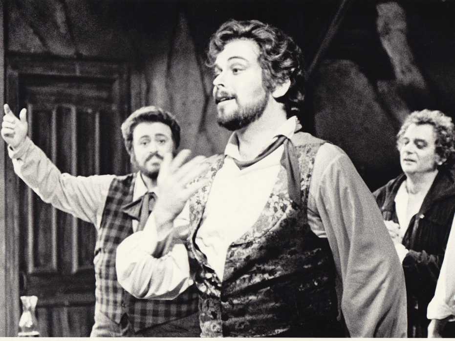 Patrick Raftery in a production of La Bohème, singing in the foreground as Luciano Pavarotti looks on in the background.