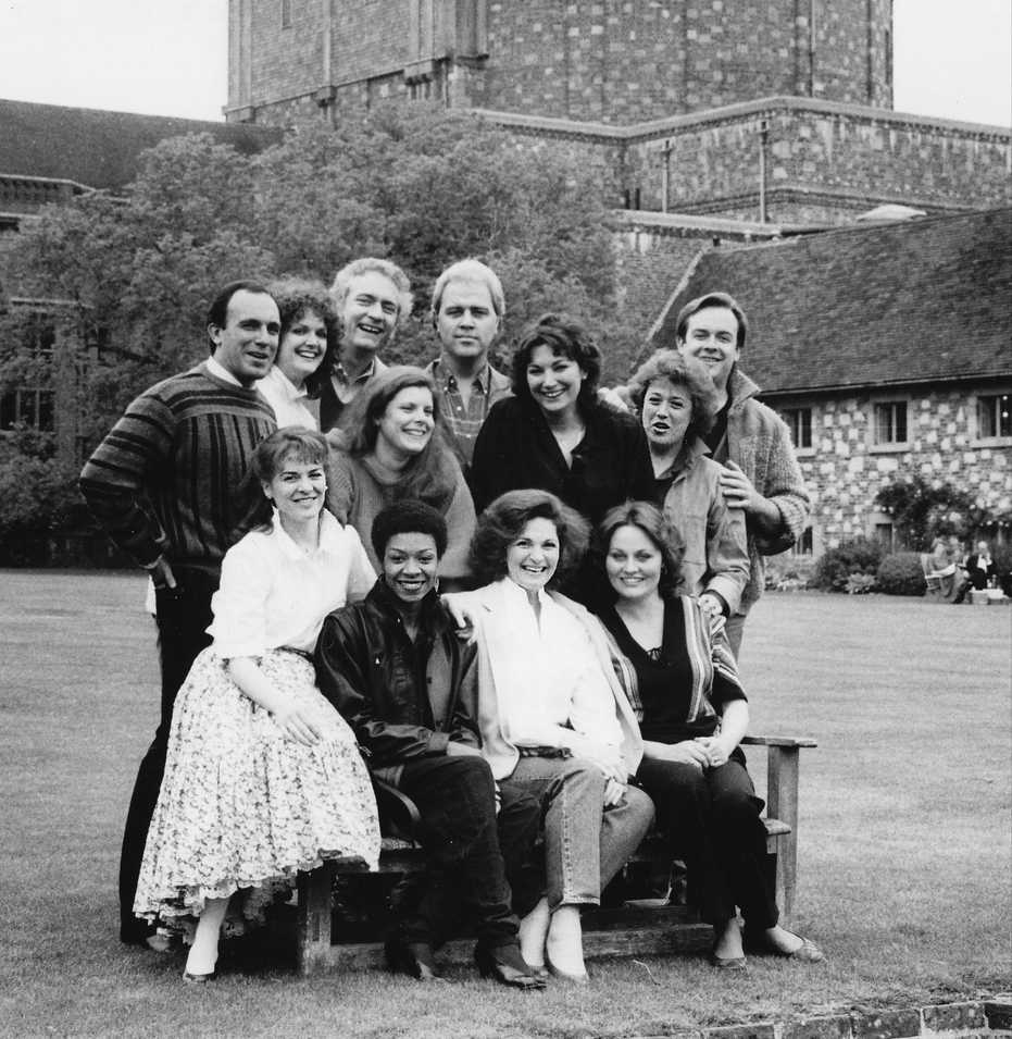 Patrick Raftery smiling with 11 other young singers in front of a school in Glyndebourne. The other singers include Dale Duesing, Ashley Putnam, Richard Stilwell, Gianna Rolandi, Dennis Bailey, Carol Vaness, Faith Esham, Cynthia Clarey, Regina Sarfaty, Delores Ziegler and Mimi Lerner.