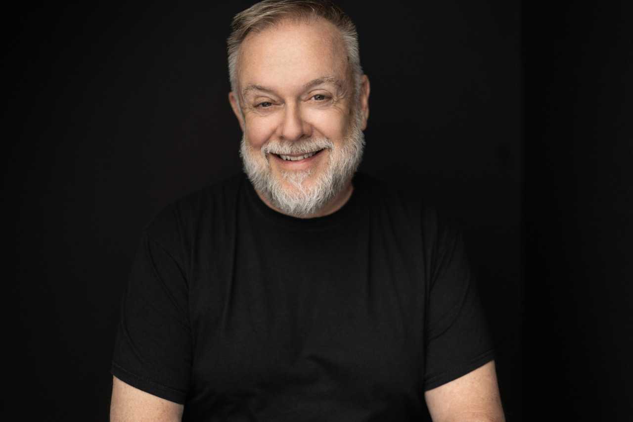 A headshot of a smiling Patrick Raftery wearing a black shirt in 2023.