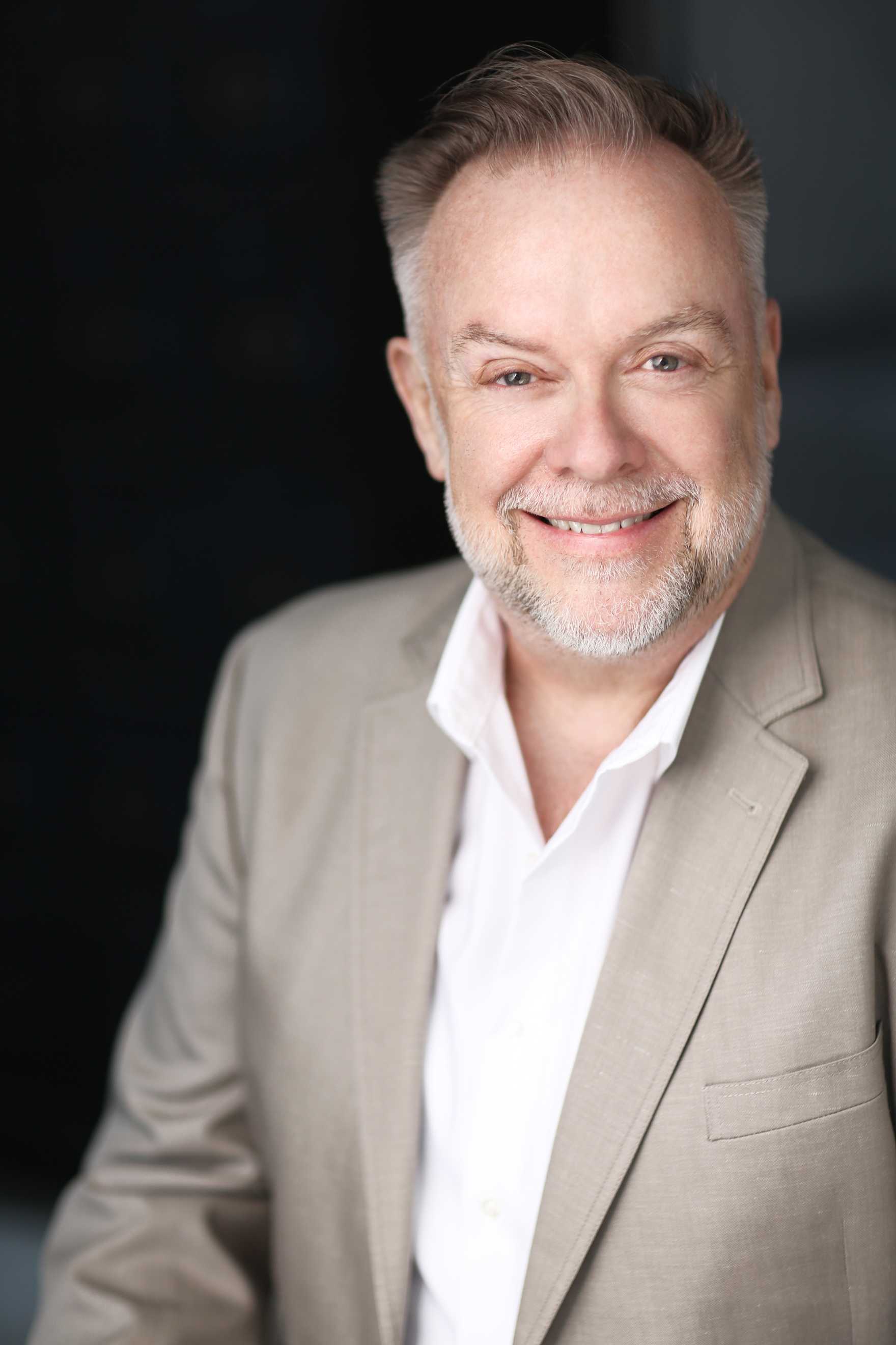 Patrick Raftery smiling broadlys and wearing a tan suit with a white shirt.