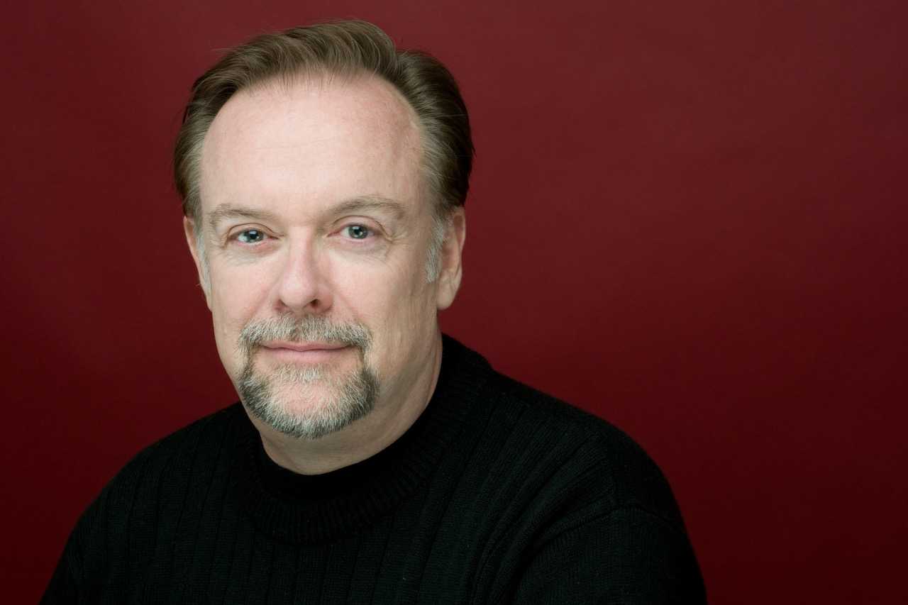 Patrick Raftery wearing a black sweater with a red wall behind him.