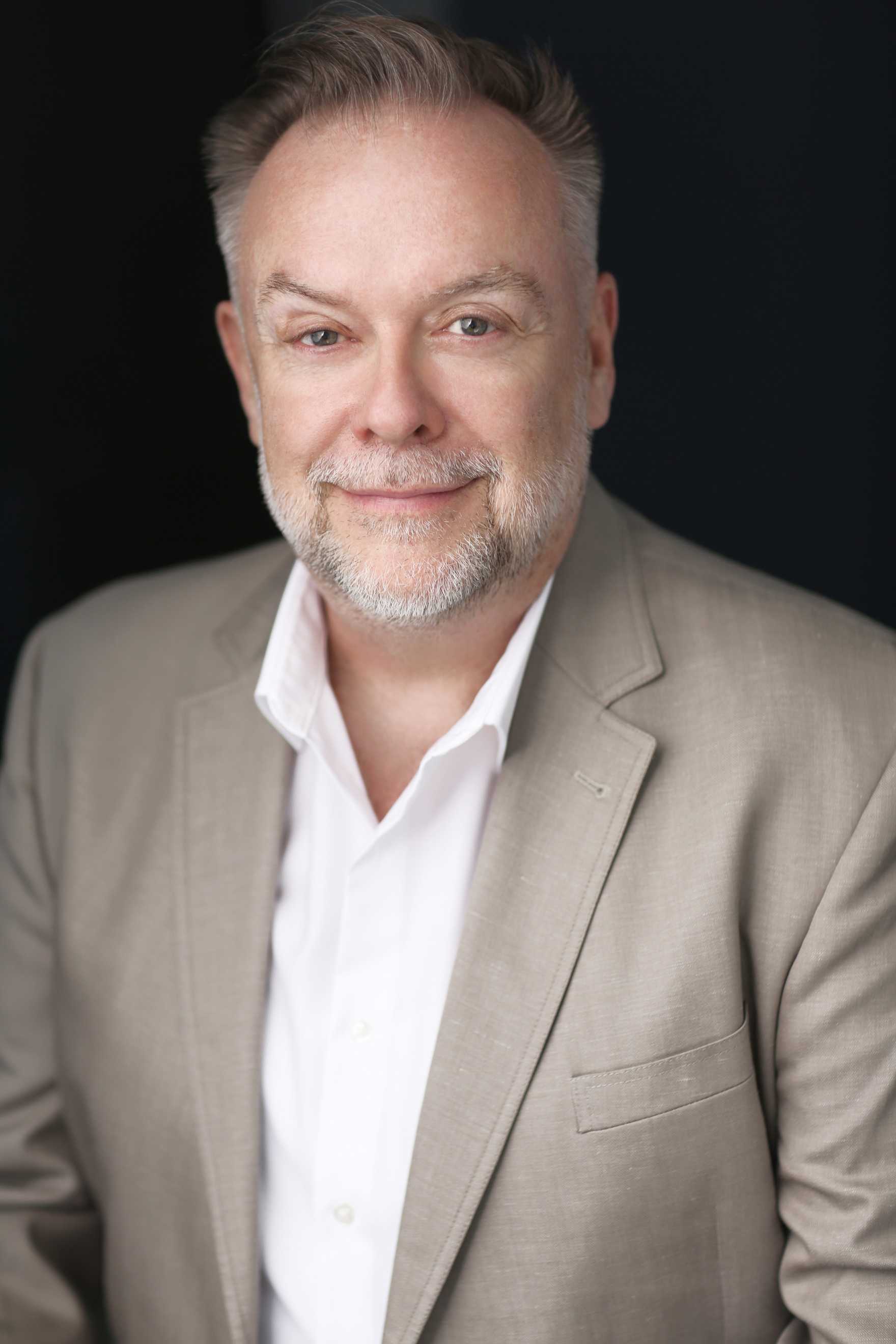 Patrick Raftery smiling subtly and wearing a tan suit with a white shirt.