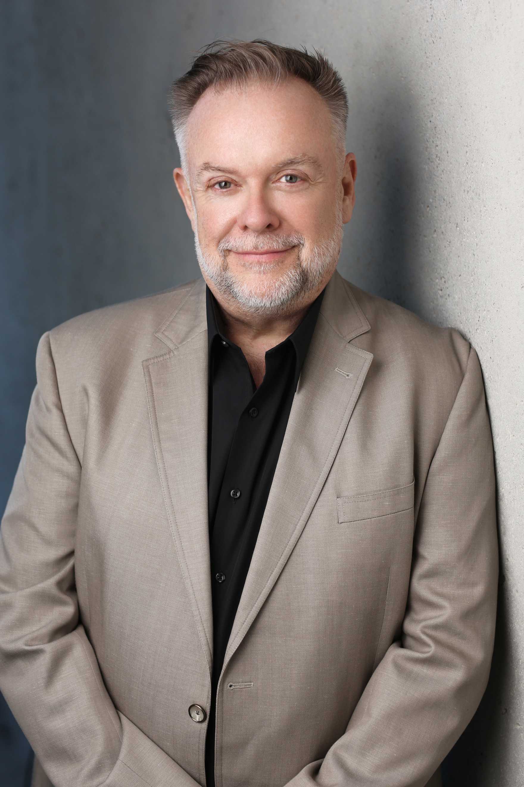 Patrick Raftery smiling and wearing a tan suit with a black shirt.