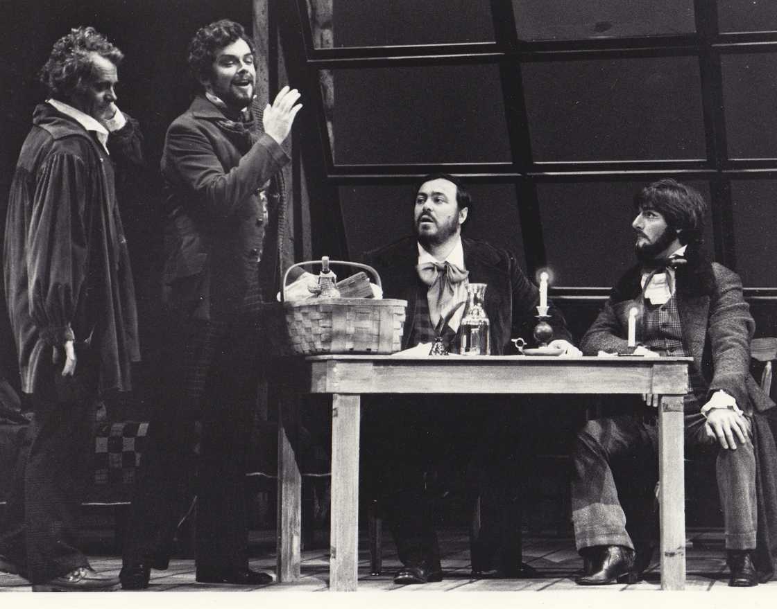 Patrick Raftery in a production of La Bohème, singing happily to two gentlemen (including Luciano Pavarotti) seated at a table.