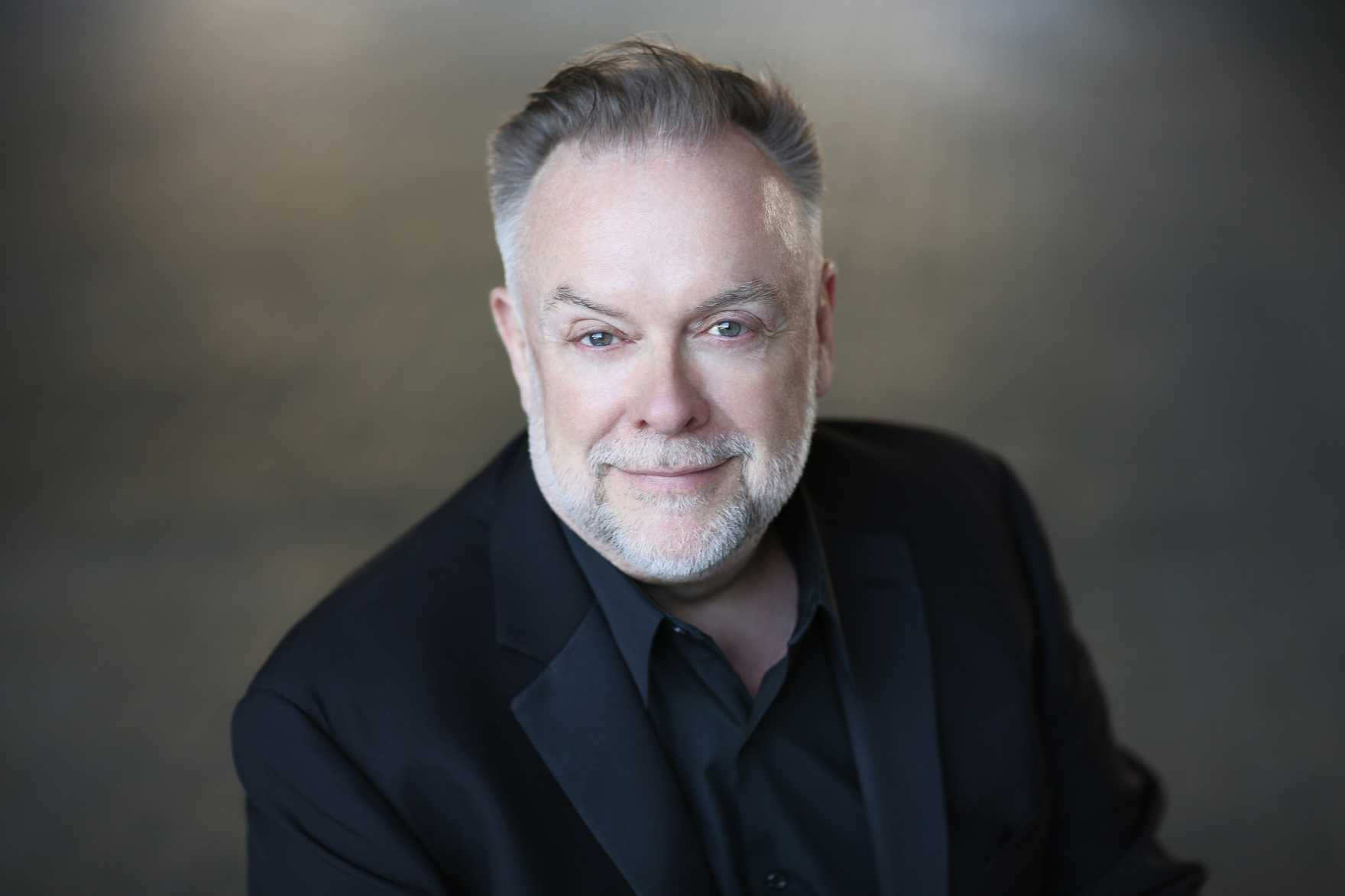 Patrick Raftery smiling and wearing a black suit with a black shirt.